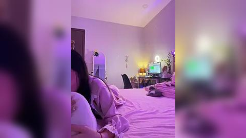 Media: A blurry video of a woman lying on a bed with a purple and pink lighted room in the background, featuring a desk, chair, and computer.