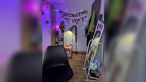 Media: Video of a dimly lit, festive room with a black chair in the foreground, \"Merry Christmas\" banner, fairy lights, and a white Christmas tree on the right.