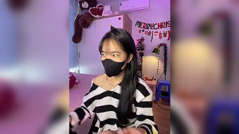 Media: Video of an Asian woman with long black hair, wearing a black face mask, black-and-white striped off-shoulder sweater, sitting indoors with a pink background, decorated with teddy bears and \"MEZOKO\" sign.