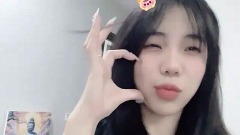 Media: Video of an East Asian woman with long black hair, fair skin, and red lipstick, holding her hands in a heart shape near her face. Background features a blurred, white wall and a framed poster.