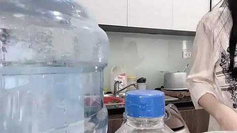 Media: Video of a woman with long black hair, wearing a white top, filling a large blue water bottle from a clear plastic jug. Kitchen background includes a sink, stove, and countertop cluttered with items.