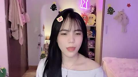 Media: Video of a young Asian woman with long black hair, wearing a white off-shoulder top, in a cozy, pink-themed room with plush toys, neon lights, and a plush bear hanging.