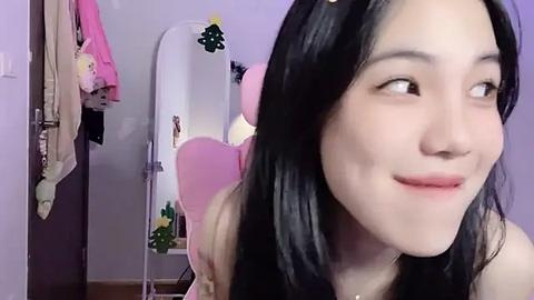 Media: Video of a young Asian woman with long black hair, wearing pink clothing, smiling. Background includes a mirror with pink heart decoration, clothes hanging, and a yellow plant.