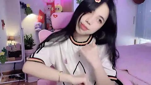 Media: Video of an Asian woman with long black hair and fair skin, wearing a white athletic shirt, seated on a pink chair in a cozy room with pastel-colored decor, including plush toys and a lamp.