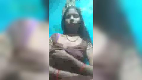 Media: Video of a woman with long hair, wearing a sari, standing in front of a turquoise background. Her face is partially blurred, and she has a necklace.