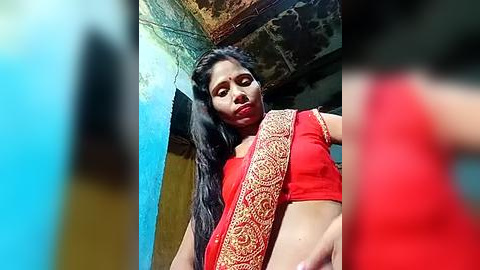 Media: Video of a woman with medium skin tone and long black hair, wearing a red sari with gold embroidery, standing in a rustic, dimly lit room with peeling paint and a blue wall.