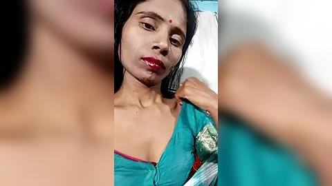 Media: Video of a South Asian woman with fair skin and long black hair, wearing a teal saree, sitting in a hospital room with a white bed. Her expression is serious and her lips are red.