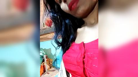 Media: A close-up video of a woman with long, dark hair, wearing a red off-shoulder top, standing indoors with a blue wall and decorative objects in the background.