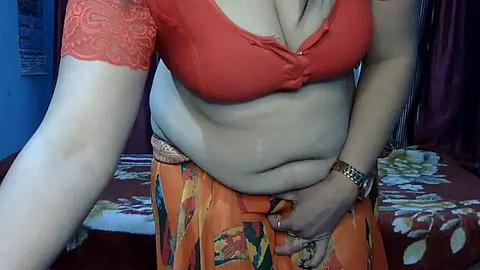 Media: Video of a plus-size woman with fair skin, wearing a red lace bra and an orange floral skirt, with a visible belly and a wristwatch, standing in a bedroom with a floral bedspread.