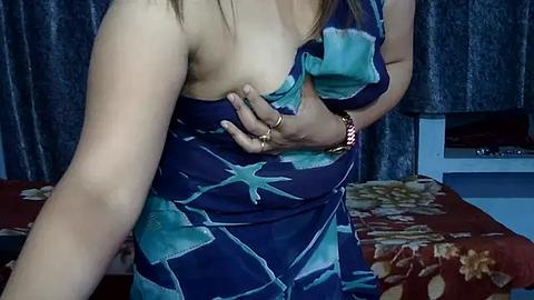 Media: Video of a woman with light skin, wearing a blue dress with teal stars, exposing her left breast, holding it with her left hand. Background shows a bed with red floral sheets, dark curtains, and a white bedside table.