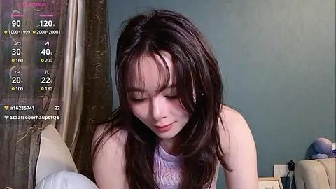 Media: Video of a young Asian woman with long dark hair, wearing a light purple sweater, sitting on a bed in a teal-colored room.