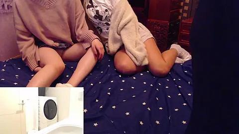 Media: Video of two young women in pajamas, one wearing a white shirt, the other in a beige sweater, kneeling on a blue bedspread with star patterns, with a close-up inset of a white toilet bowl in the bottom left corner.