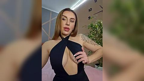 Media: Video of a confident, light-skinned woman with long, straight brown hair, wearing a black halterneck dress with a plunging neckline, revealing ample cleavage. She has a tattoo on her left arm and is indoors, with a blurred background of greenery and a clock.