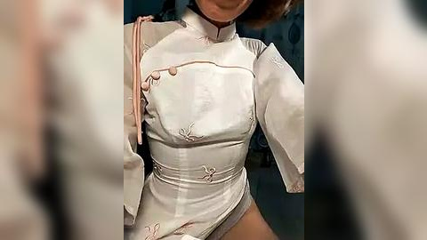 Media: A video of a woman in a white, high-collared cheongsam dress with pink floral embroidery, worn with a pink sash, standing against a blurred background.