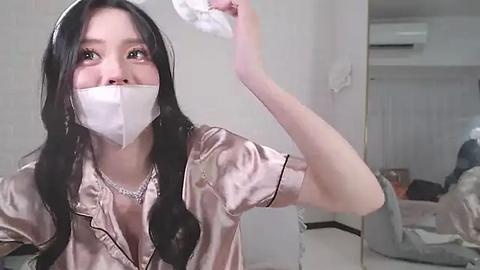 Media: Video of a young East Asian woman with long black hair, wearing a pink satin robe, white mask, and a silver necklace, cleaning a mirror in a modern, minimalistic bedroom with white walls and light furniture.