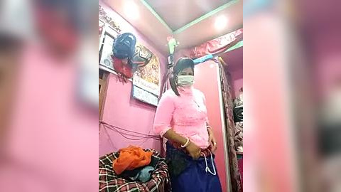 Media: Video of a woman in a pink shirt and blue skirt, wearing a face mask, standing in a small, cluttered room with pink walls and hanging bags, a mirror, and a plaid chair.