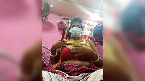 Media: Video of a woman in a pink room, wearing a red bra, yellow top, and face mask, adjusting her bra, with clothing and a fan visible in the background.