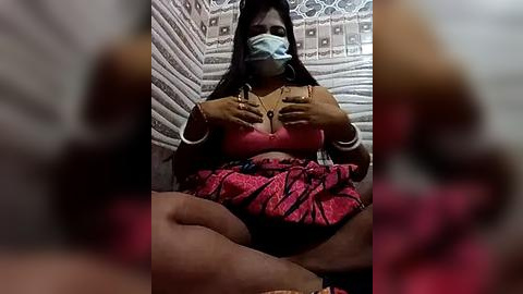 Media: Video of a dark-skinned woman with long black hair, wearing a pink floral dress, sitting cross-legged on a bed. She covers her breasts with her hands, wearing a surgical mask. Background features a patterned wall.