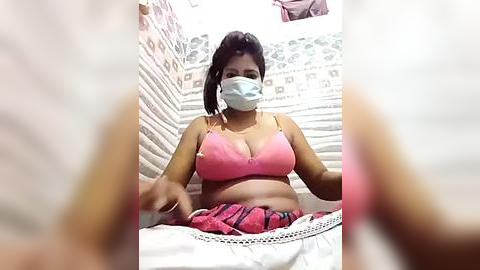 Media: A video of a plus-sized woman with dark skin, wearing a pink bra and face mask, sitting on a bed with white sheets, in a cluttered room with patterned wallpaper.