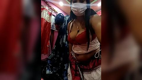 Media: A video of a young woman with medium skin tone and long black hair wearing a red bra, white mask, and a red and white skirt in a cluttered, dimly lit room with hanging clothes.