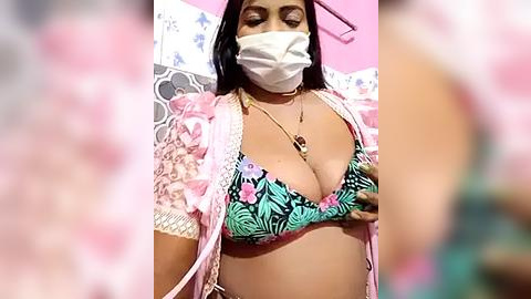 Media: A video of a woman with light brown skin, wearing a pink face mask, a black-and-green bikini top, and a sheer pink kimono. She has medium-sized breasts, long black hair, and is standing in a pink room with floral wallpaper.