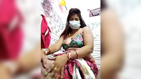 Media: Video of a South Asian woman with medium skin tone, wearing a colorful floral bikini top, face mask, and red sari, sitting in a tiled bathroom, with a blurred figure in the foreground.