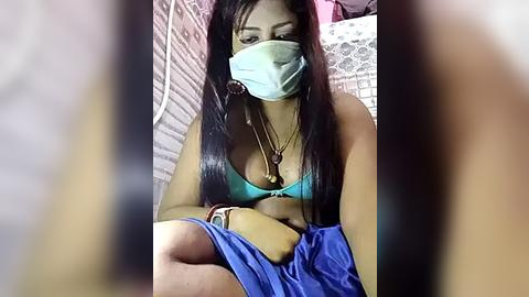 Media: Video of a woman with medium brown skin, long black hair, wearing a turquoise bra, blue pants, and a blue surgical mask, lying on a bed with white and patterned pillows in a dimly lit room.