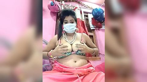 Media: Video of a South Asian woman with medium skin tone, wearing a pink sari, a white face mask, and a string of pearls, covering her breasts with her hands, in a pink bedroom with colorful decorations and balloons.