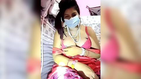 Media: A video of an Asian woman with medium skin tone, wearing a pink bra, blue face mask, pearl necklace, and red skirt, sitting on a bed with a pink wall in the background.