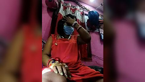 Media: Video of a woman in a red saree, wearing a blue face mask, sitting on a bed, with a pink wall and hanging clothes in the background.