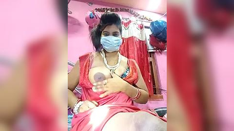 Media: A video of a South Asian woman in a pink dress, covering her breast with her hand, wearing a mask, in a cluttered room with red clothes hanging.