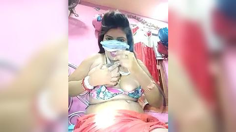 Media: Video of a young woman with a mask, wearing a bikini top and shorts, sitting on a pink bed in a cluttered room.