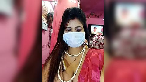 Media: Video of a woman with long dark hair, wearing a blue face mask, pearl necklace, and red saree, seated in a pink room with a TV in the background.