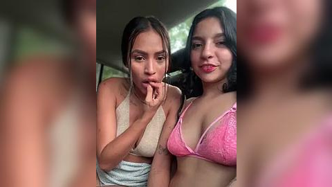 Media: Video of two young Latina women, one in a beige tank top, the other in a pink lace bra, smiling intimately indoors.