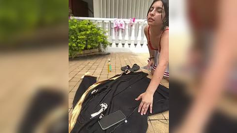 Media: Video of a woman in a striped tank top kneeling beside a black bag with a white phone on a brick patio, surrounded by greenery and a white railing.