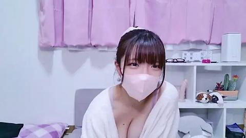 Media: Video of an East Asian woman with a white mask, wearing a white bathrobe, sitting on a bed with pink curtains, white shelves with cactus and stuffed animals in the background.