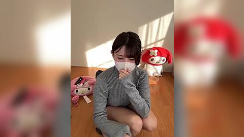 Media: Video of a young Asian woman with short black hair, wearing a gray sweater and a white mask, sitting on a wooden floor, surrounded by plush toys in a bright room.