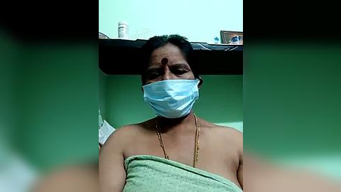 Media: Video of a woman with a light brown complexion, wearing a light green towel and a blue medical mask, sitting indoors with a plain green wall in the background.