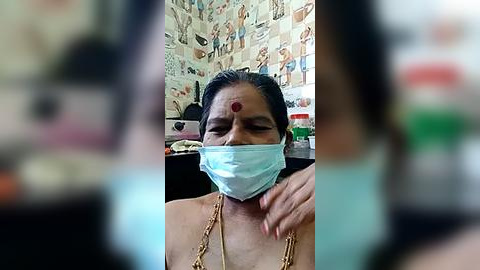 Media: Video of a South Asian woman wearing a blue surgical mask, a red bindi on her forehead, and a gold chain necklace, seated indoors with wallpaper depicting Hindu deities in the background.