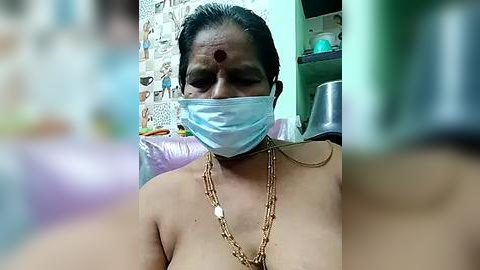 Media: A video of a topless woman with medium skin tone and short black hair, wearing a blue surgical mask, a gold necklace, and a red bindi. The background shows a kitchen with a tiled wall, a metal pot, and a green cabinet.