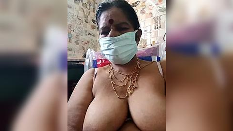 Media: A video of a South Asian woman with a medium skin tone, wearing a light blue surgical mask, revealing large breasts. She is seated indoors, surrounded by cluttered shelves.