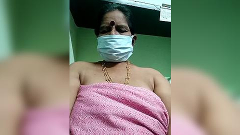 Media: Video of an Indian woman with medium skin tone, wearing a pink textured cloth, a gold chain, and a face mask, in a green-walled room with a black ceiling.