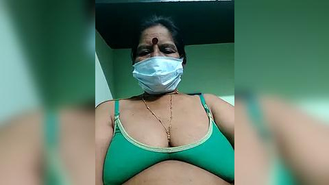 Media: Video of a South Asian woman with medium brown skin, wearing a green bra, white mask, and a bindi, with blurred green walls in the background.