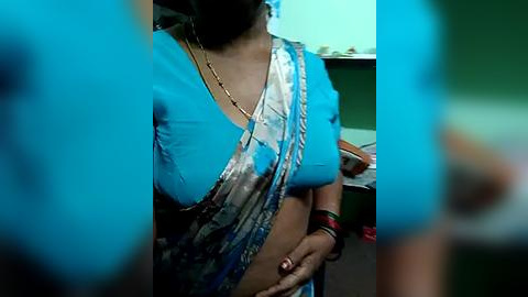 Media: Video of a woman in a blue sari with silver trim, wearing a gold necklace and bracelets, standing indoors with blurred background.