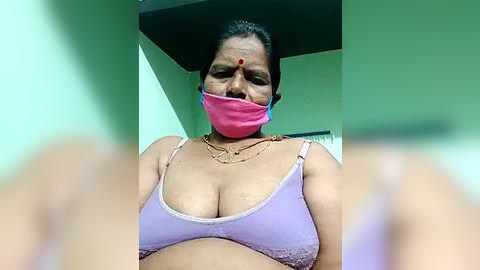 Media: Video of an Indian woman with medium skin tone, wearing a pink mask, purple bra, and gold necklace, indoors with green walls.