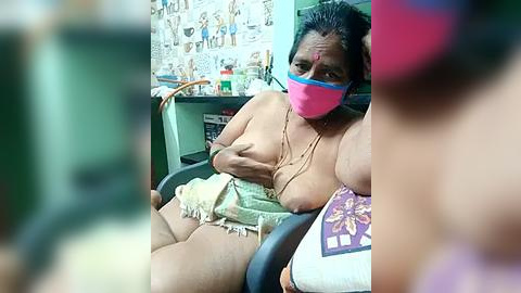 Media: A video of an overweight, middle-aged woman with dark skin and dark hair, wearing a pink mask, sitting topless on a chair in a cluttered room with posters on the wall.