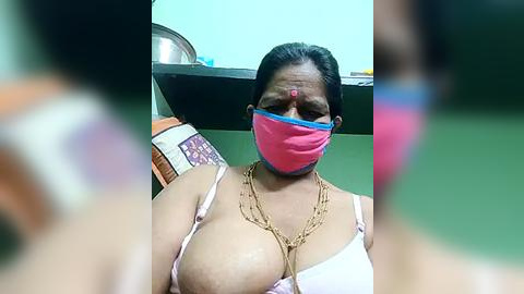 Media: A video of an Indian woman with a medium complexion, wearing a pink mask, a white bra, and gold necklaces, seated on a cushioned chair with a blurred background.