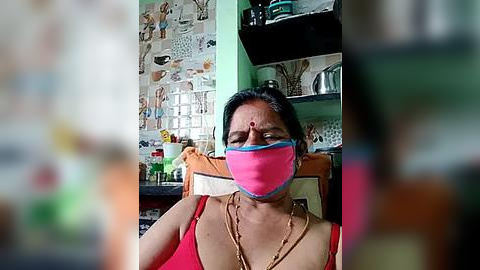 Media: Video of an Indian woman in a pink mask and red tank top, sitting in a cluttered kitchen with a colorful tiled wall and a sink.