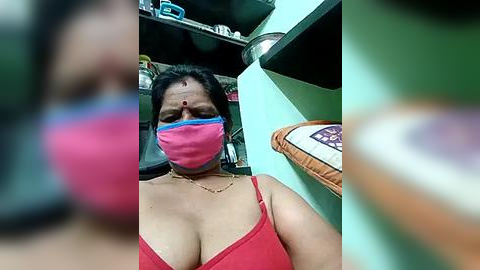 Media: Video of an Indian woman with medium skin tone, wearing a pink mask, red bra, and a gold necklace. She's in a cluttered room with shelves and a green wall, blurred background.