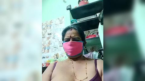 Media: Video of an elderly Indian woman with medium-dark skin tone, wearing a pink mask, purple sleeveless top, and gold necklace, sitting in a cluttered room with a green wall and shelves filled with household items.